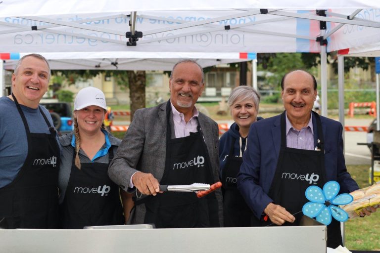 MoveUP leadership (Brian Martens, Christy Slusarenko and Annette Toth) stand with Minister Harry Bains and Speaker Raj Chouhan underneath a MoveUP tent behind a barbecue grill wearing MoveUP aprons