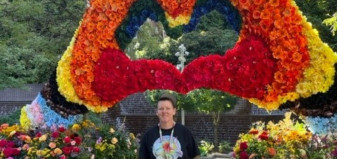 Person poses in front of floral arrangement made to look like two hands making the shape of a heart